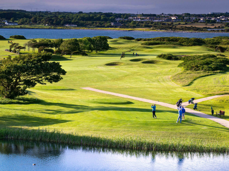 A scenic view of Galway Bay Golf Resort, showcasing a lush green fairway with golfers in action. The course is surrounded by natural dunes, trees, and water features, with the stunning backdrop of Galway Bay and a distant shoreline under a partly cloudy sky.