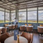 Bright restaurant lounge with large windows overlooking a golf course and bay, featuring blue banquette seating, tan leather chairs, small gold lamps on tables, and a server arranging place settings.