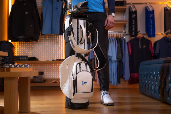 A person standing in a pro shop beside a white Galway Bay–branded golf bag with quilted detailing. The bag is upright on the floor, surrounded by racks of golf apparel and accessories. The person is wearing golf shoes and dark pants, with only their lower body visible.