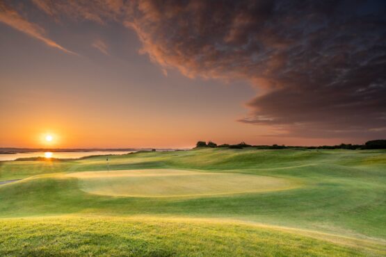 A golf green overlooking a calm body of water at sunset, with warm orange light on the horizon and dramatic clouds sweeping across the sky.