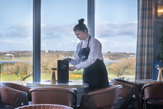 Restaurant-4 A waitress in a Galway Bay Golf uniform is setting a table in a beautifully designed dining area. She is arranging a branded menu on a round table, which is accompanied by stylish chairs and elegant gold-accented table decor. Large windows in the background offer a stunning view of the golf course and surrounding landscape under a partly cloudy sky.