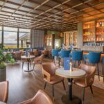 A beautifully designed bar and lounge area at Galway Bay Golf, featuring a warm, inviting atmosphere with a mix of plush leather chairs and blue barstools. The wooden coffered ceiling adds character, while large windows offer stunning views of the golf course and coastline. A well-stocked backlit bar showcases a selection of premium spirits, beers, and wines. Thoughtful decor elements, including greenery, elegant lighting, and neatly arranged tables with gold candle holders, create a refined yet relaxed setting—perfect for unwinding after a round of golf or enjoying a casual drink with friends.