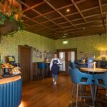 A stylish café and lounge area at Galway Bay Golf, featuring a warm and inviting ambiance. The space is adorned with floral-patterned wallpaper, a coffered wooden ceiling, and cozy lighting. A barista station with a green Astoria coffee machine and a selection of freshly baked pastries sits in the foreground. Plush blue barstools surround high-top tables, each set with elegant gold candle holders and branded menus. A friendly staff member in a navy apron walks through the room, adding to the welcoming atmosphere. Framed photos decorate the walls, creating a sense of heritage and charm in this refined yet relaxed setting.