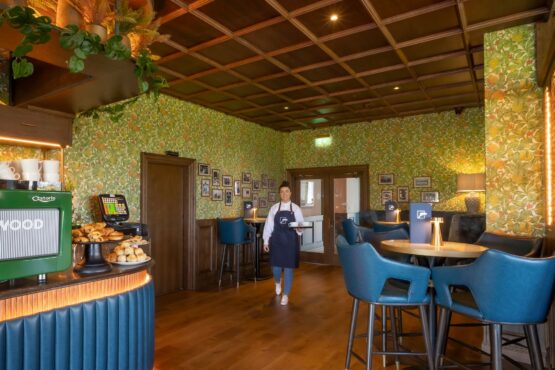 entry-2iron A stylish café and lounge area at Galway Bay Golf, featuring a warm and inviting ambiance. The space is adorned with floral-patterned wallpaper, a coffered wooden ceiling, and cozy lighting. A barista station with a green Astoria coffee machine and a selection of freshly baked pastries sits in the foreground. Plush blue barstools surround high-top tables, each set with elegant gold candle holders and branded menus. A friendly staff member in a navy apron walks through the room, adding to the welcoming atmosphere. Framed photos decorate the walls, creating a sense of heritage and charm in this refined yet relaxed setting.