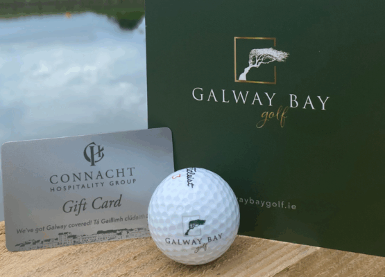 gbg-vouchers-1200x628 Close-up of a Galway Bay Golf gift card, a Connacht Hospitality Group gift card, and a Galway Bay–branded golf ball arranged on a wooden railing with a calm lake and grassy landscape blurred in the background.