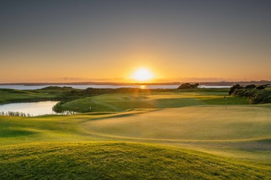 golf-section Golden sunset over a beautifully maintained golf course beside a calm body of water. The green and fairway are softly illuminated by the setting sun, casting long shadows across the landscape and reflecting warm light on the horizon.