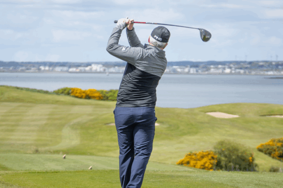 A golfer at Galway Bay Golf follows through on a powerful drive, watching the ball soar down the fairway. Dressed in a black cap and jacket, he stands against a stunning backdrop of rolling greens, golden gorse bushes, and the calm waters of Galway Bay. The bright sky and distant cityscape add to the picturesque setting, capturing the essence of a perfect day on the course.