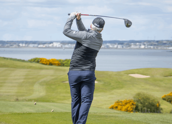 A golfer at Galway Bay Golf follows through on a powerful drive, watching the ball soar down the fairway. Dressed in a black cap and jacket, he stands against a stunning backdrop of rolling greens, golden gorse bushes, and the calm waters of Galway Bay. The bright sky and distant cityscape add to the picturesque setting, capturing the essence of a perfect day on the course.
