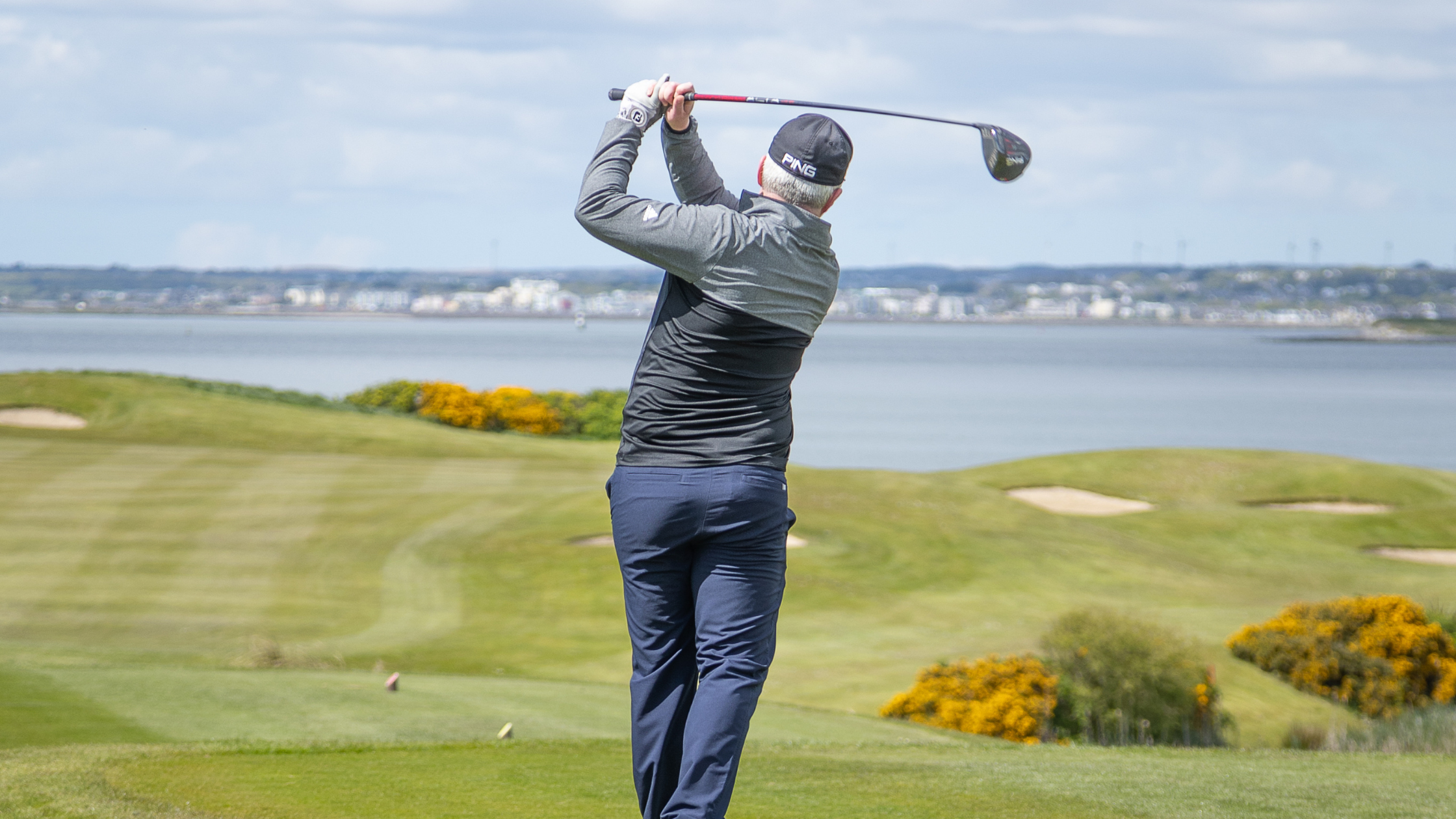 A golfer at Galway Bay Golf follows through on a powerful drive, watching the ball soar down the fairway. Dressed in a black cap and jacket, he stands against a stunning backdrop of rolling greens, golden gorse bushes, and the calm waters of Galway Bay. The bright sky and distant cityscape add to the picturesque setting, capturing the essence of a perfect day on the course.