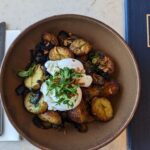 A rustic brunch dish served at Galway Bay Golf, featuring golden roasted baby potatoes with a crispy exterior, topped with perfectly poached eggs, fresh herbs, and crispy shallots. The dish is presented in an earthy brown bowl, highlighting the rich colors and textures. A Galway Bay Golf menu with gold embossing is placed beside the bowl, complementing the elegant yet cozy dining setup. Silverware and a napkin are neatly arranged, ready for a delicious and hearty meal.