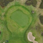 A stunning aerial view of a green at Galway Bay Golf, surrounded by natural rough and a single, strategically placed sand bunker. The well-manicured putting surface contrasts beautifully with the surrounding fairway and rugged terrain. A golf cart is parked near the edge of the hole, adding a sense of scale to this picturesque and challenging layout.