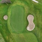 A stunning aerial view of a green at Galway Bay Golf, surrounded by two strategically placed sand bunkers and framed by lush fairways. The smooth putting surface contrasts beautifully with the natural landscape, featuring a nearby water hazard and windswept trees. The well-designed layout presents both a scenic and challenging approach for golfers.