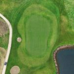 A stunning aerial view of a green at Galway Bay Golf, bordered by two sand bunkers and a scenic water hazard. The well-manicured putting surface contrasts beautifully with the surrounding fairway and rugged natural edges. A golf cart is parked along the winding pathway, adding scale to this picturesque and challenging hole.