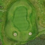 A stunning aerial view of a green at Galway Bay Golf, featuring two strategically placed sand bunkers that add both challenge and visual appeal. The well-manicured putting surface is seamlessly integrated into the surrounding fairway, with natural rough and a nearby water hazard enhancing the scenic beauty of the hole.
