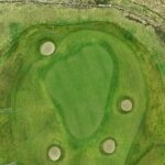A stunning aerial view of a green at Galway Bay Golf, surrounded by four strategically placed sand bunkers. The well-manicured putting surface contrasts beautifully with the surrounding fairway and natural rough. A low stone wall borders the hole, blending seamlessly into the rugged coastal landscape, adding both character and challenge to this picturesque setting.