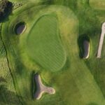 A stunning aerial view of a green at Galway Bay Golf, surrounded by three strategically placed sand bunkers and bordered by natural rough. The fairway’s smooth contours blend seamlessly with the rugged coastal landscape. A golf cart is parked near the pathway, casting a long shadow in the soft, golden light.