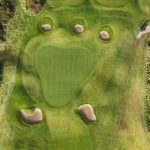 A stunning aerial view of a green at Galway Bay Golf, surrounded by five strategically placed sand bunkers that add both challenge and beauty to the hole. The fairway seamlessly blends with the surrounding natural landscape, featuring rolling terrain and patches of rough. A golf cart is parked near the pathway, casting a long shadow in the soft, golden light.