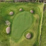 A stunning aerial view of a green at Galway Bay Golf, surrounded by five strategically placed sand bunkers that enhance both the challenge and beauty of the hole. The putting surface is smoothly contoured, blending seamlessly with the surrounding fairway and natural rough. A lone tree stands near one of the bunkers, adding a scenic touch to the carefully designed landscape.