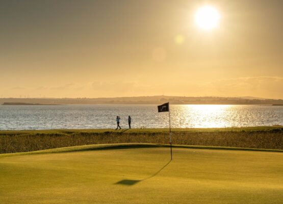 A stunning sunset view at Galway Bay Golf Resort, with the sun casting a golden glow over the putting green and shimmering waters of Galway Bay. A flag bearing the resort's logo stands gently in the breeze, while one golfer takes a shot and another observes near the shoreline in the distance, adding to the serene and picturesque atmosphere.
