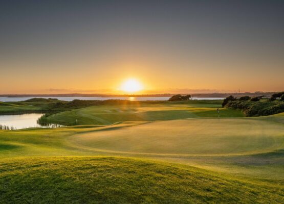 A breathtaking sunset over Galway Bay Golf, casting a golden glow across the immaculate green and surrounding fairways. The flagstick stands tall against the backdrop of calm waters and rolling dunes, creating a serene yet dramatic scene. The combination of soft light, long shadows, and natural beauty captures the essence of coastal links golf at its finest.