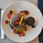 A beautifully presented stack of fluffy pancakes served at Galway Bay Golf, topped with fresh berries, orange segments, and a dusting of powdered sugar. The pancakes are arranged on a textured white plate, accompanied by a small pot of rich chocolate sauce for drizzling. A navy blue Galway Bay Golf menu with gold embossing is placed beside the plate, adding a touch of elegance. Silverware and a neatly folded napkin complete the setting, making for a delicious and picturesque breakfast by the bay.