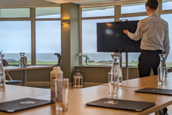 A modern conference room at Galway Bay Golf, where a professionally dressed individual is presenting on a large screen TV. The room features floor-to-ceiling windows offering stunning views of the golf course and the Atlantic Ocean, creating an inspiring setting for meetings. The wooden tables are neatly arranged with branded folders, glass pitchers of water, and drinking glasses. Decorative elements like a lantern and floral arrangements add a touch of elegance. The setup is ideal for corporate events, strategy sessions, or executive retreats, blending business with breathtaking coastal scenery.
