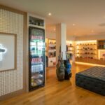 The stylish and well-lit pro shop at Galway Bay Golf, featuring a modern design with wooden flooring and illuminated display shelves. Golf apparel, accessories, and equipment are neatly arranged, with a selection of golf bags standing near a refrigerated drinks display. A plush tufted bench sits in the center, and a TV screen in the background shows a golf tournament. The resort’s signature tree logo is elegantly backlit on the wall.