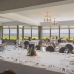 A beautifully arranged dining space at Galway Bay Golf, set for a special event. The room features elegantly dressed tables with crisp white linens, fine glassware, polished silverware, and chic gold candle holders. Stylish round-backed chairs complement the sophisticated setting. Large panoramic windows offer stunning views of the lush green golf course, golden gorse, and the sparkling waters of Galway Bay. Soft natural light enhances the refined ambiance, while contemporary chandeliers add a warm glow, making it a perfect venue for weddings, celebrations, or fine dining experiences.