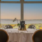 An elegant dining setup at Galway Bay Golf, featuring a round table draped in a crisp white tablecloth, set with fine glassware, polished silverware, and a stylish gold candle holder. A delicate orchid centerpiece adds a touch of sophistication. Large bay windows frame a breathtaking view of the golf course, with rolling greens, golden gorse, and the shimmering waters of Galway Bay stretching into the horizon. The warm hues of the evening sky enhance the tranquil and luxurious atmosphere, making it an ideal spot for a memorable dining experience.
