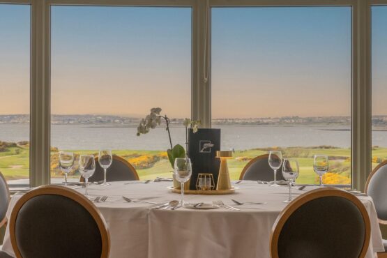 restaurant-view An elegant dining setup at Galway Bay Golf, featuring a round table draped in a crisp white tablecloth, set with fine glassware, polished silverware, and a stylish gold candle holder. A delicate orchid centerpiece adds a touch of sophistication. Large bay windows frame a breathtaking view of the golf course, with rolling greens, golden gorse, and the shimmering waters of Galway Bay stretching into the horizon. The warm hues of the evening sky enhance the tranquil and luxurious atmosphere, making it an ideal spot for a memorable dining experience.