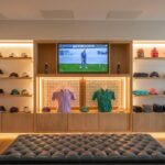 The pro shop at Galway Bay Golf, featuring a stylish and modern design with neatly displayed golf apparel, including hats, shirts, and accessories. Two mannequin torsos showcase polo shirts in purple and green, illuminated by elegant backlighting. A large TV screen above the display is broadcasting a golf tournament. A plush, tufted leather bench sits in the center, inviting guests to relax while browsing. The shop’s sleek wooden shelves and warm lighting create an inviting atmosphere for golfers looking to update their gear in style.