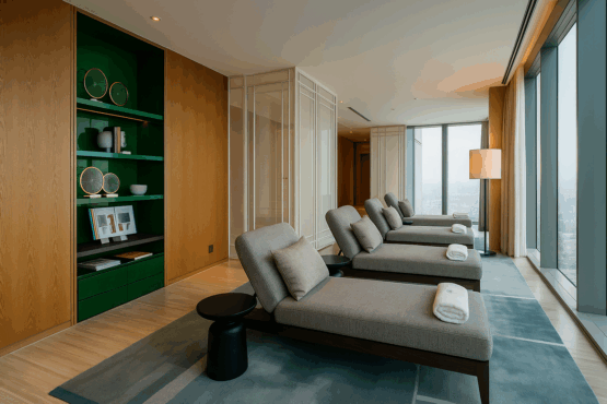 th-relaxation-room4 Modern relaxation lounge with large windows overlooking a clandscape, featuring four cushioned chaise lounges each with a towel and pillow, a floor lamp, and built-in wooden shelving with decorative accents.