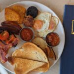 A hearty Full Irish Breakfast served at Galway Bay Golf, beautifully presented on a white plate. The meal includes crispy rashers of bacon, golden hash browns, fried eggs with slightly runny yolks, grilled sausages, black and white pudding, and roasted tomatoes. Accompanying the dish are two small pots of baked beans and a rich tomato relish. Generous slices of toasted bread complete the plate. A glass of fresh orange juice sits nearby, alongside a navy blue Galway Bay Golf menu with gold embossing, adding a touch of elegance to the dining experience.