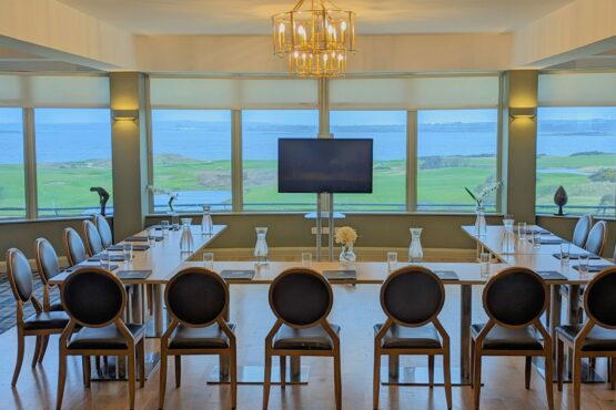 A sophisticated conference room at Galway Bay Golf, set up in a U-shaped meeting arrangement with elegant wooden chairs and a sleek modern table. A large screen TV is positioned at the front for presentations, framed by floor-to-ceiling windows that showcase breathtaking views of the golf course and Galway Bay. The room is decorated with minimalist floral arrangements and features glass pitchers of water and neatly arranged glasses, adding a refined touch. A gold chandelier hangs from the ceiling, enhancing the room's stylish and professional ambiance—perfect for corporate meetings, strategy sessions, or executive retreats.